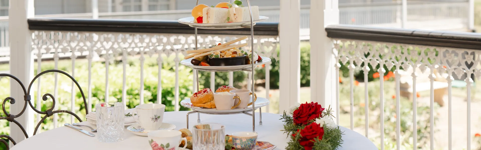 High tea setup with tiered tray, pastries, sandwiches, and tea cups on a veranda.
